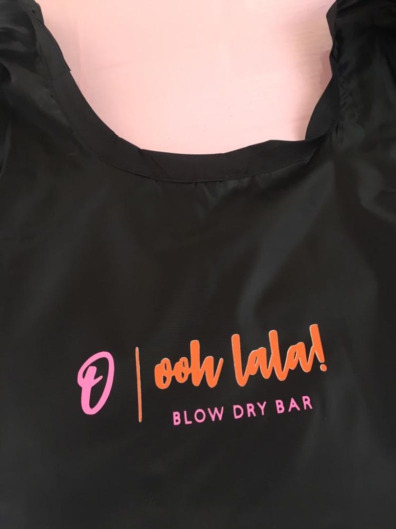 K&ouml;nnte beinhalten: Schwarze wiederverwendbare Einkaufstasche mit dem Text "ooh la la!" und "BLOW DRY BAR" in orangefarbener und rosafarbener Schrift.