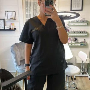 May include: A woman wearing a black uniform with the text "ML'S BEAUTY" embroidered on the chest. She is standing in a room with a white wall and a mirror behind her.