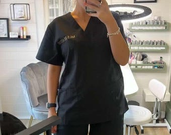 May include: A woman wearing a black uniform with the text "ML'S BEAUTY" embroidered on the chest. She is standing in a room with a white wall and a mirror behind her.