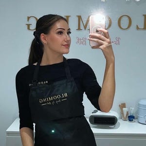 K&ouml;nnte beinhalten: Eine Frau tr&auml;gt eine schwarze Sch&uuml;rze mit dem Text "BLOOMING Beauty Bar" in goldenen Buchstaben. Sie macht ein Selfie in einem Salon.