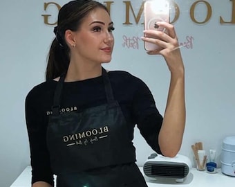 May include: A woman wearing a black apron with the text "BLOOMING Beauty Bar" in gold lettering. She is taking a selfie in a salon setting.