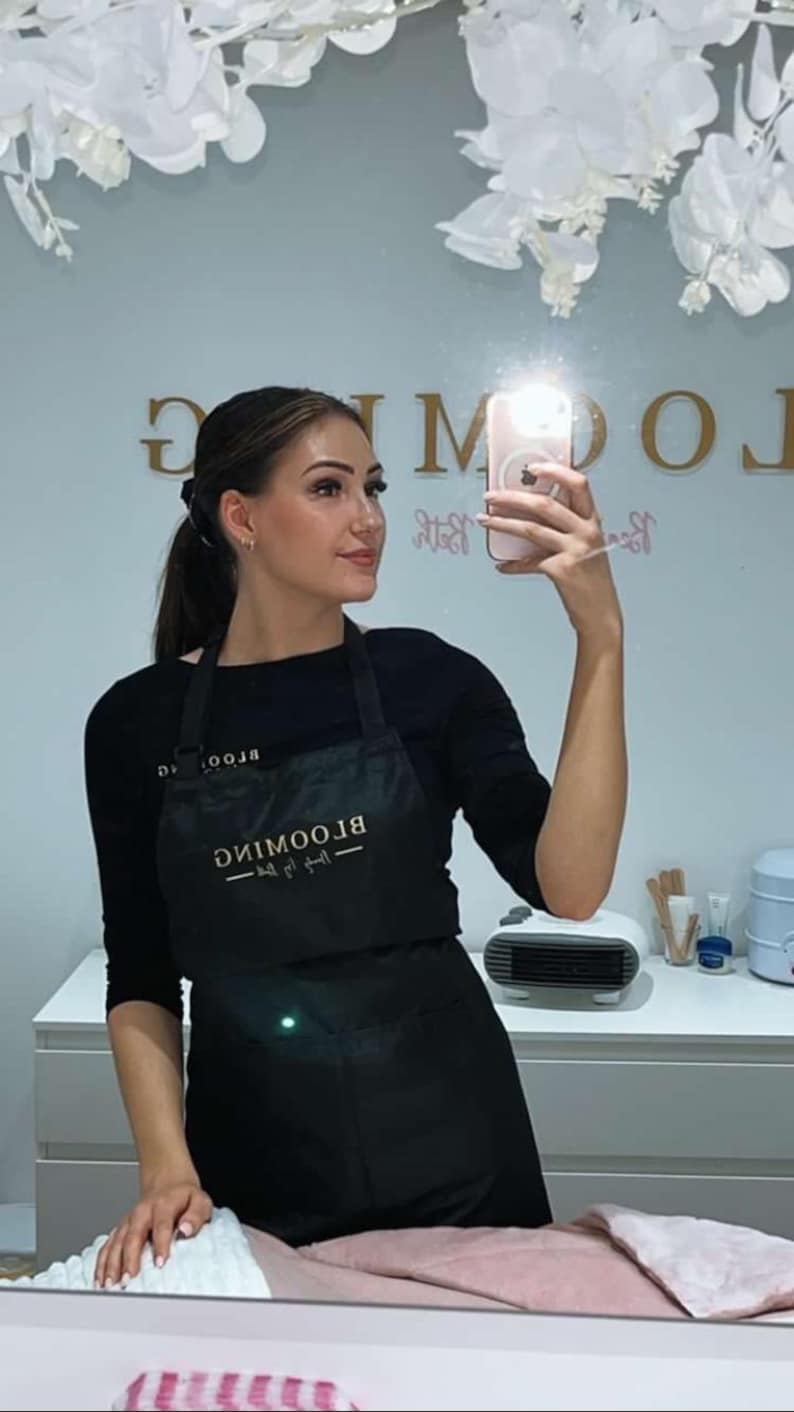 K&ouml;nnte beinhalten: Eine Frau tr&auml;gt eine schwarze Sch&uuml;rze mit dem Text "BLOOMING Beauty Bar" in goldenen Buchstaben. Sie macht ein Selfie in einem Salon.