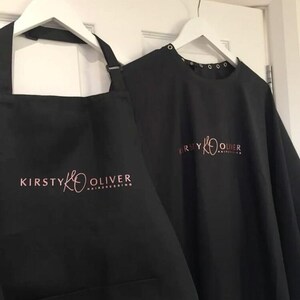 K&ouml;nnte beinhalten: Schwarze Friseur-Sch&uuml;rze und -Umhang mit dem Logo "Kirsty K Oliver Hairdressing" in rosa Schrift.