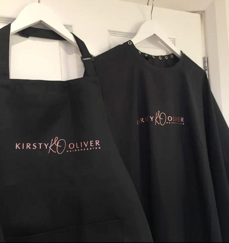 K&ouml;nnte beinhalten: Schwarze Friseur-Sch&uuml;rze und -Umhang mit dem Logo "Kirsty K Oliver Hairdressing" in rosa Schrift.