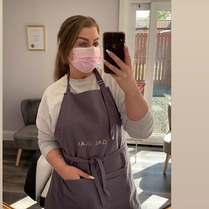 K&ouml;nnte beinhalten: Eine Frau tr&auml;gt eine graue Sch&uuml;rze mit dem gestickten Text "GAL GLAM" auf der Vorderseite. Sie tr&auml;gt eine rosa Gesichtsmaske und macht ein Selfie in einem Spiegel.