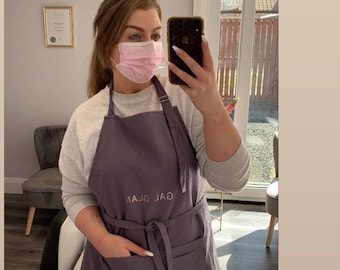 May include: A woman wearing a gray apron with the text "GAL GLAM" embroidered on the front. She is wearing a pink face mask and taking a selfie in a mirror.