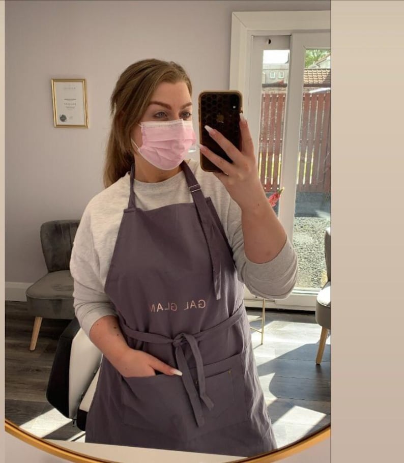 K&ouml;nnte beinhalten: Eine Frau tr&auml;gt eine graue Sch&uuml;rze mit dem gestickten Text "GAL GLAM" auf der Vorderseite. Sie tr&auml;gt eine rosa Gesichtsmaske und macht ein Selfie in einem Spiegel.