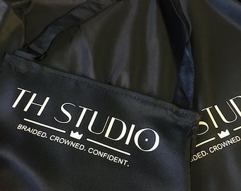 May include: Black salon cape with the text "TH STUDIO" in white, along with the words "BRAIDED. CROWNED. CONFIDENT." The cape has a pink lining and an adjustable neck strap.