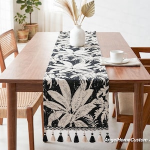 Può includere: Runner da tavolo bianco e nero con motivo a foglie. Il runner è decorato con nappe nere e crema. È posto su un tavolo di legno con un vaso bianco e una tazza e un piattino.