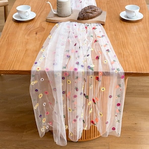 May include: A white, sheer table runner adorned with embroidered flowers in various colours. The runner is laid on a wooden table, with a loaf of bread, cups, and a teapot visible. The floral embroidery includes red, yellow, and purple flowers.