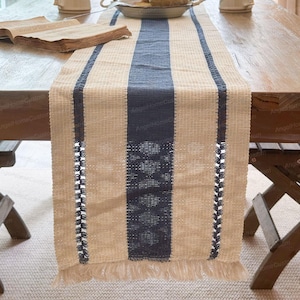 May include: A beige and navy blue table runner with a woven texture. The runner features a central navy blue stripe and two thinner navy blue stripes. The edges are finished with a fringe.