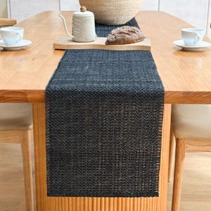 Peut inclure: Un chemin de table tissé gris foncé avec un motif texturé, placé sur une table en bois clair. Le chemin de table est centré, avec une théière, du pain et des tasses visibles. La table est entourée de chaises en bois, créant un cadre de salle à manger chaleureux.