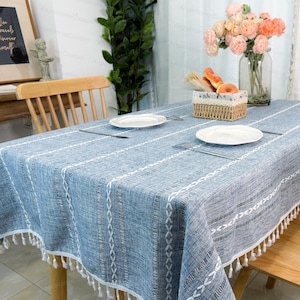 May include: A blue and white striped tablecloth with fringe trim. The tablecloth is made of a linen blend and has a subtle woven texture.