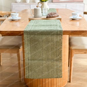 Puede incluir: Un camino de mesa verde tejido está centrado en una mesa de madera, con una tetera, pan y tazas de té. El camino de mesa tiene una apariencia texturizada y agrega un elemento decorativo al comedor. La mesa está rodeada de sillas.