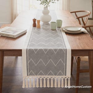 May include: A rectangular table runner featuring a grey and white chevron pattern with a geometric herringbone design. The runner is finished with long, cream-coloured fringe tassels on both ends, creating a textured border along the table surface.