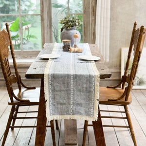 Puede incluir: Una mesa de madera rústica con dos sillas, puesta con un camino de mesa a rayas azul y blanco, platos blancos y saleros y pimenteros. El camino de mesa tiene un borde de flecos.