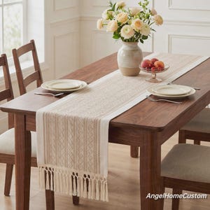 May include: A long rectangular table runner featuring a cream and tan geometric woven pattern with long fringe tassels on the ends. The runner is placed on a dark wood dining table with white plates, a glass bowl of fruit, and a vase of flowers.