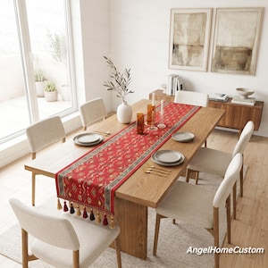Op de afbeelding: Een rood en goudkleurig tafelloper met kwastjes, gepresenteerd op een houten eettafel. De loper heeft een geometrisch ontwerp en wordt geflankeerd door crèmekleurige stoelen. De tafel is gedekt met borden en goudkleurig bestek.
