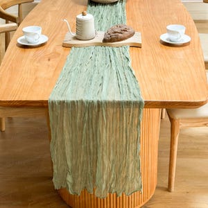 Puede incluir: Un camino de mesa arrugado verde claro sobre una mesa de madera. La mesa está decorada con tazas y platillos blancos, una tetera y un pan. La mesa tiene un acabado de madera clara.