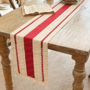 Puede incluir: Camino de mesa rectangular con rayas verticales rojas y crema. El corredor está hecho de tela tejida y tiene un borde con flecos. Un libro abierto descansa sobre la mesa de madera.