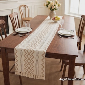 May include: A long, cream-colored table runner featuring a repeating brown zigzag and diamond pattern with vertical textured stripes. The runner has fringe trim on the ends and is placed down the center of a dark brown wooden dining table.
