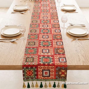 Könnte beinhalten: Ein langer, rechteckiger Tischläufer mit einem sich wiederholenden geometrischen Patchwork-Muster in Rot-, Grün-, Braun- und Cremetönen. Der Tischläufer ist am unteren Rand mit dekorativen Quasten in Grün und Gold versehen.