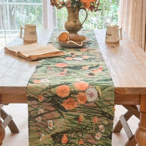 Puede incluir: Una mesa de madera está decorada con un camino de mesa floral verde. El camino de mesa presenta flores naranjas y blancas sobre un fondo verde. Un jarrón de flores, dos tazas, un plato de pasteles y un libro abierto están sobre la mesa.