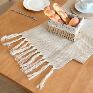 May include: A cream-coloured woven table runner with white tassels. A basket of bread sits on the runner, next to a white plate and a cup. The table is made of light brown wood.