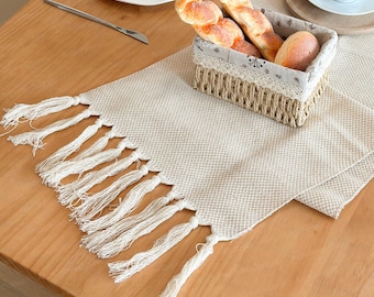 May include: A beige woven table runner with white fringe detail. A basket filled with bread sits on the runner, alongside a white plate and a cup. The table is made of light brown wood.
