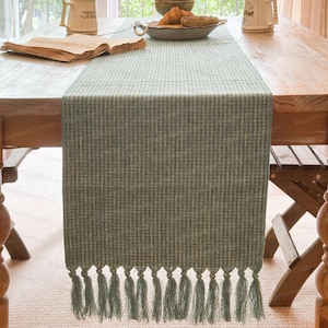Puede incluir: Camino de mesa verde salvia con tejido texturizado y borlas decorativas. El camino de mesa está colocado sobre una mesa de madera, añadiendo un toque de elegancia rústica. El fleco del camino de mesa añade un elemento decorativo.