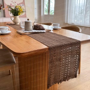 Op de afbeelding: Een bruine geweven tafelloper op een houten tafel. De loper heeft een textuur en is geplaatst over een lichtbruine tafel. Een theepot, kopjes en een brood zitten ook op de tafel.