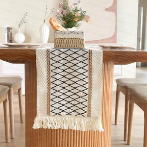 Könnte beinhalten: Ein weißer und schwarzer geometrischer Tischläufer mit Fransenbordüre. Der Läufer liegt auf einem Holztisch mit zwei Stühlen.