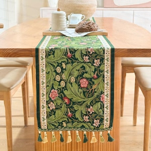 Puede incluir: Camino de mesa floral verde con un patrón detallado de hojas y flores en tonos verdes, rosas y crema. El camino de mesa tiene un borde crema y verde y borlas en la parte inferior. Colocado sobre una mesa de madera.