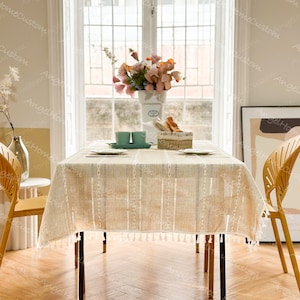 May include: A beige tablecloth with white fringe and a striped pattern. The tablecloth is laid on a wooden table with two chairs. There are flowers in a vase on the table.