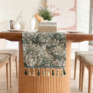 Könnte beinhalten: Ein floraler Tischläufer mit grün-weißem Muster, verziert mit Quasten. Er liegt auf einem Holztisch, ergänzt durch beige Stühle. Ein Korb mit Brot und Blumen ergänzt die Tischdekoration.