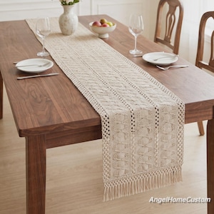 May include: A long, rectangular beige table runner featuring an intricate knit pattern with vertical textured columns and a decorative fringe along the bottom edge. The runner is laid flat across a dark brown wooden dining table with place settings.