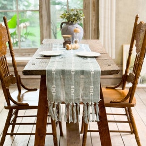 May include: A rustic wooden dining table set with a sage green and white patterned table runner. Two wooden chairs with carved backs flank the table. A vase with greenery, plates, and a pepper grinder are on the table.