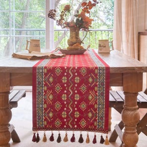 Può includere: Una tovaglia rossa con un motivo geometrico e nappe. La tovaglia è posta su un tavolo di legno, con un vaso di fiori e tazze visibili. Il design della tovaglia include motivi a rombi e zigzag nei toni del verde, giallo e blu.