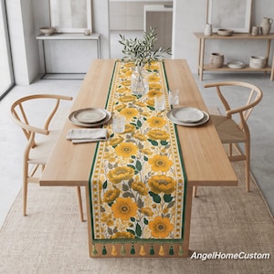 May include: A long rectangular table runner featuring a vibrant floral pattern of yellow flowers and green leaves on a cream background, bordered by dark green stripes and finished with alternating yellow and green tassels at both ends.