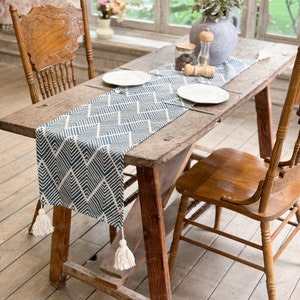 Op de afbeelding: Een blauwe en witte geometrische tafelrunner met witte kwastjes op een rustieke houten tafel met twee houten stoelen.