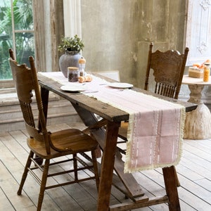 Puede incluir: Un camino de mesa rosa y blanco con borde de flecos se encuentra sobre una mesa de madera rústica con dos sillas de madera. El camino de mesa está decorado con un sutil patrón de líneas blancas.