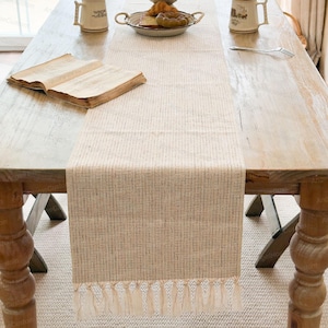 Op de afbeelding: Een beige tafelloper met een textuurpatroon en franjedetails, geplaatst op een houten tafel. Een open boek, een bord met eten en twee mokken staan ook op de tafel. De tafel heeft een lichte houten afwerking.