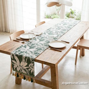 Puede incluir: Una mesa de comedor de madera con un camino de mesa floral verde y blanco. El camino de mesa tiene una franja con borlas amarillas, verdes y beige. Platos blancos, vasos y cubiertos están sobre la mesa.