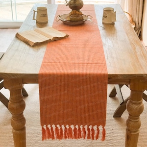 Puede incluir: Un camino de mesa tejido naranja con flecos, colocado sobre una mesa de madera. El camino de mesa está centrado, con un libro y tazas a cada lado. Una bandeja decorativa y un jarrón también están sobre la mesa.