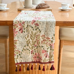 May include: A floral table runner with a cream background and a pattern of pink flowers and green leaves. The runner has a burgundy border with tassels. The runner is approximately 60 inches long.