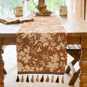 Puede incluir: Camino de mesa floral marrón y crema con flecos de borlas. El patrón floral repite hojas y flores. El camino de mesa está colocado sobre una mesa de madera, con un libro y tazas visibles.