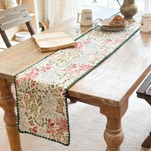 May include: A cream-coloured table runner featuring pink and green floral patterns. The runner, edged with green, is laid on a wooden table. A book, mugs, and a plate of food are also visible on the table.