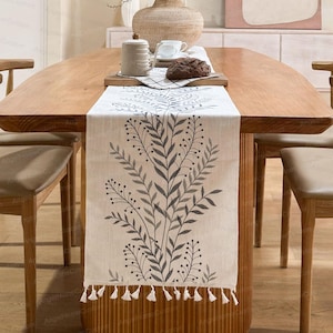 以下が含まれることがあります： 葉やベリーを描いたグレーのボタニカルデザインのクリーム色のテーブルランナー。ランナーの下部には白いタッセルのフリンジが付いています。ランナーは木製のテーブルの上に置かれています。