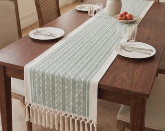 May include: A long rectangular table runner featuring a sage green and cream woven geometric pattern with vertical stripes and diamond motifs. The runner is finished with long cream-colored fringe tassels on both ends. The text AngelHomeCustom is visible.