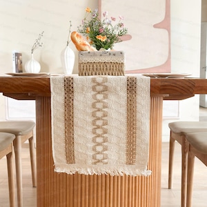 Könnte beinhalten: Ein beigefarbener und brauner gewebter Tischläufer mit geometrischem Design, Fransen und vertikalen Streifen. Der Läufer wird auf einem Holztisch präsentiert, ergänzt durch einen Korb mit Blumen und Brot, was eine rustikale Essästhetik schafft.